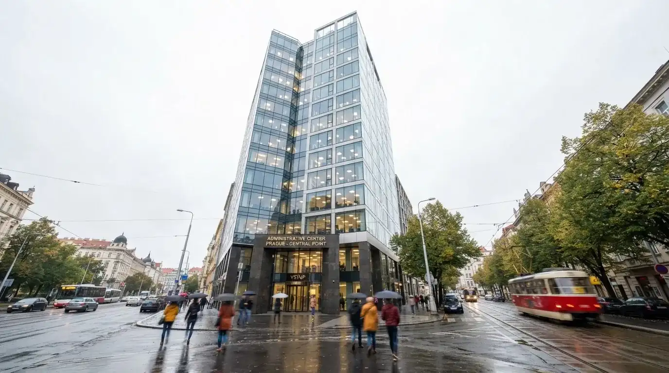 Fasáda moderní administrativní budovy s skleněnými okny odráží ranní oblohu nad rušnou pražskou ulicí.